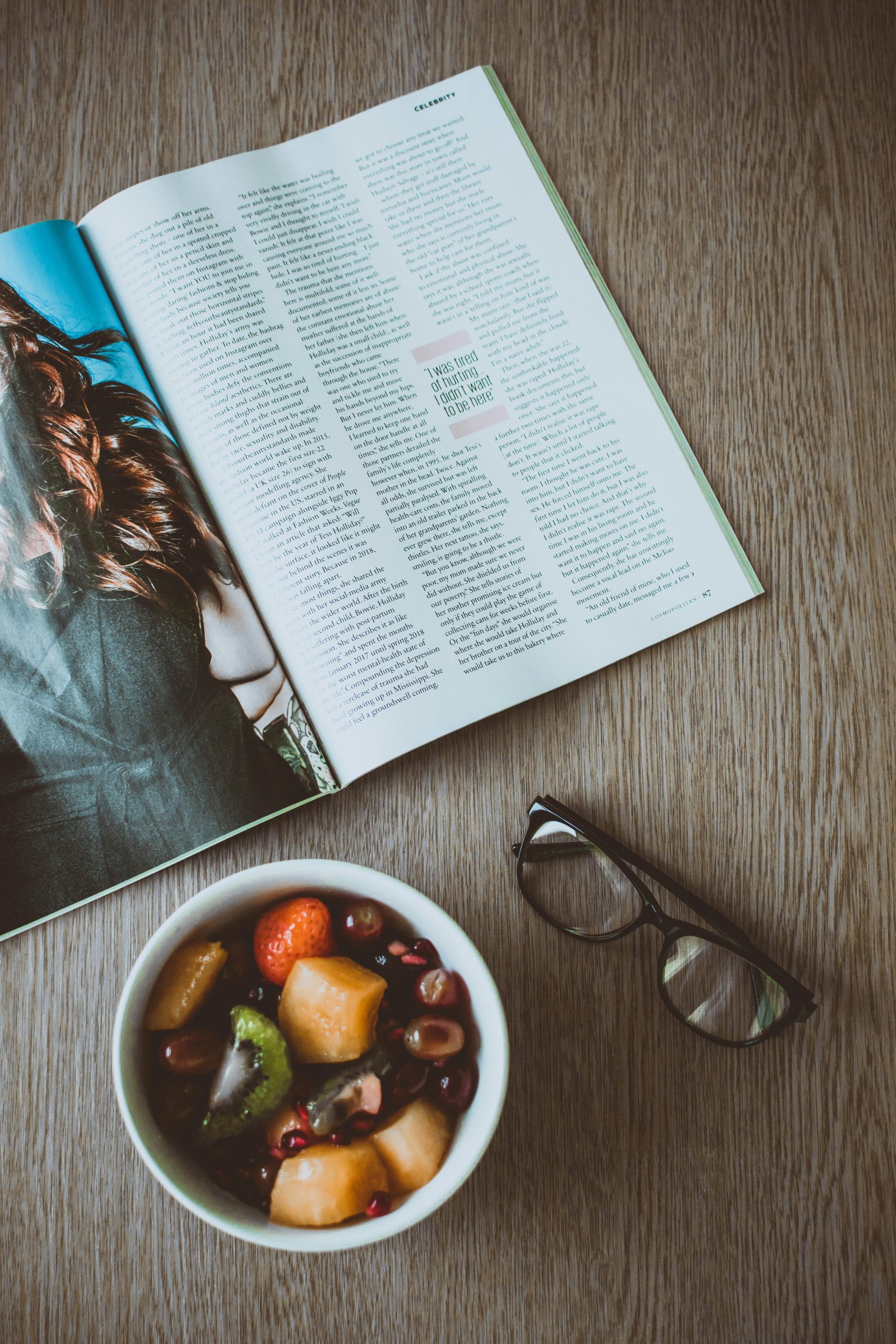 A fresh fruit bowl, open magazine, and glasses on a wooden table, perfect for lifestyle and food content.